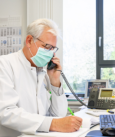 Werkarzt Dr. Wolfgang Großwieser sitzt an einem Schreibtisch und telefoniert. Er trägt dabei einen Mund-Nasen-Schutz. (Foto)